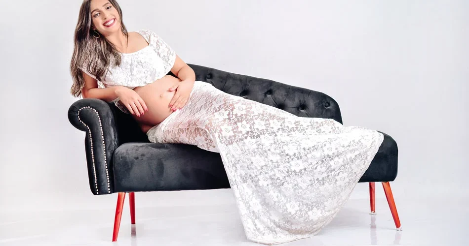 Ensaio de gestante pousando com um vestido branco e no sofá preto.